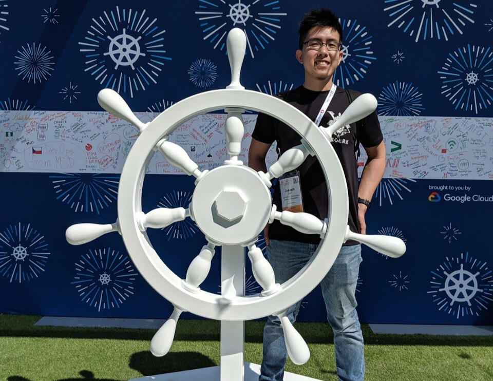 Me at KubeCon 2019 in Barcelona standing behind a giant white Kubernetes wheel!